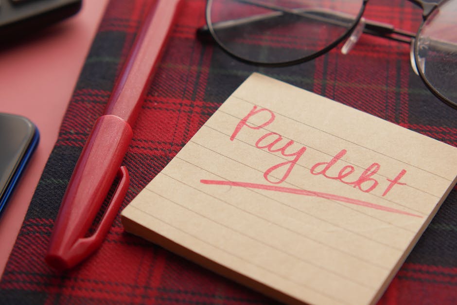 Close-up of a note reading  Pay debt  next to a red pen on a plaid fabric, emphasizing financial reminders.